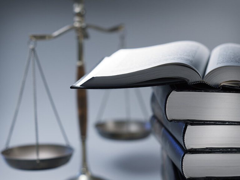 A stack of law books stands in front of a justice scale that is slightly out of focus.  On top of the stack is an open law book.