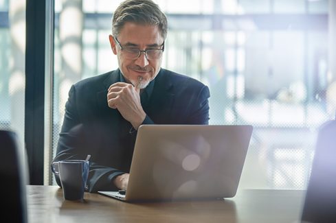 Business portrait - businessman sitting in in office working with laptop computer. Mature age, middle age, mid adult man in 50s with happy confident smile. Copy space.