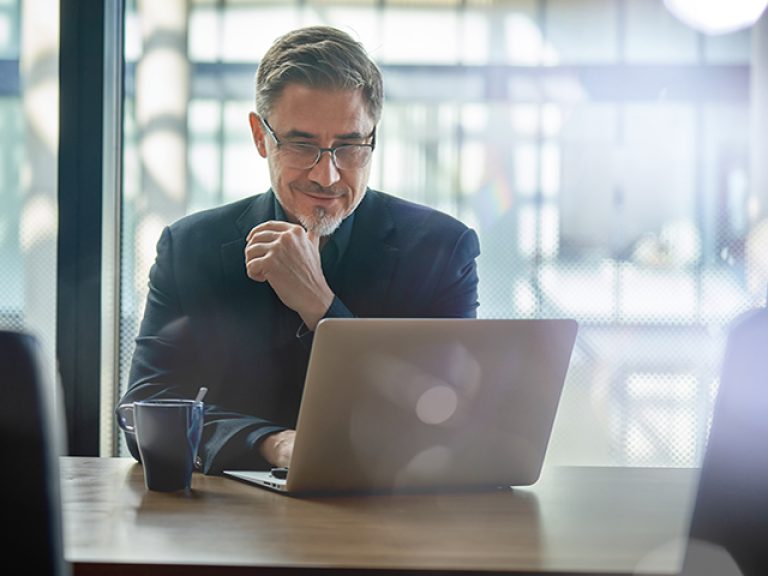 Business portrait - businessman sitting in in office working with laptop computer. Mature age, middle age, mid adult man in 50s with happy confident smile. Copy space.